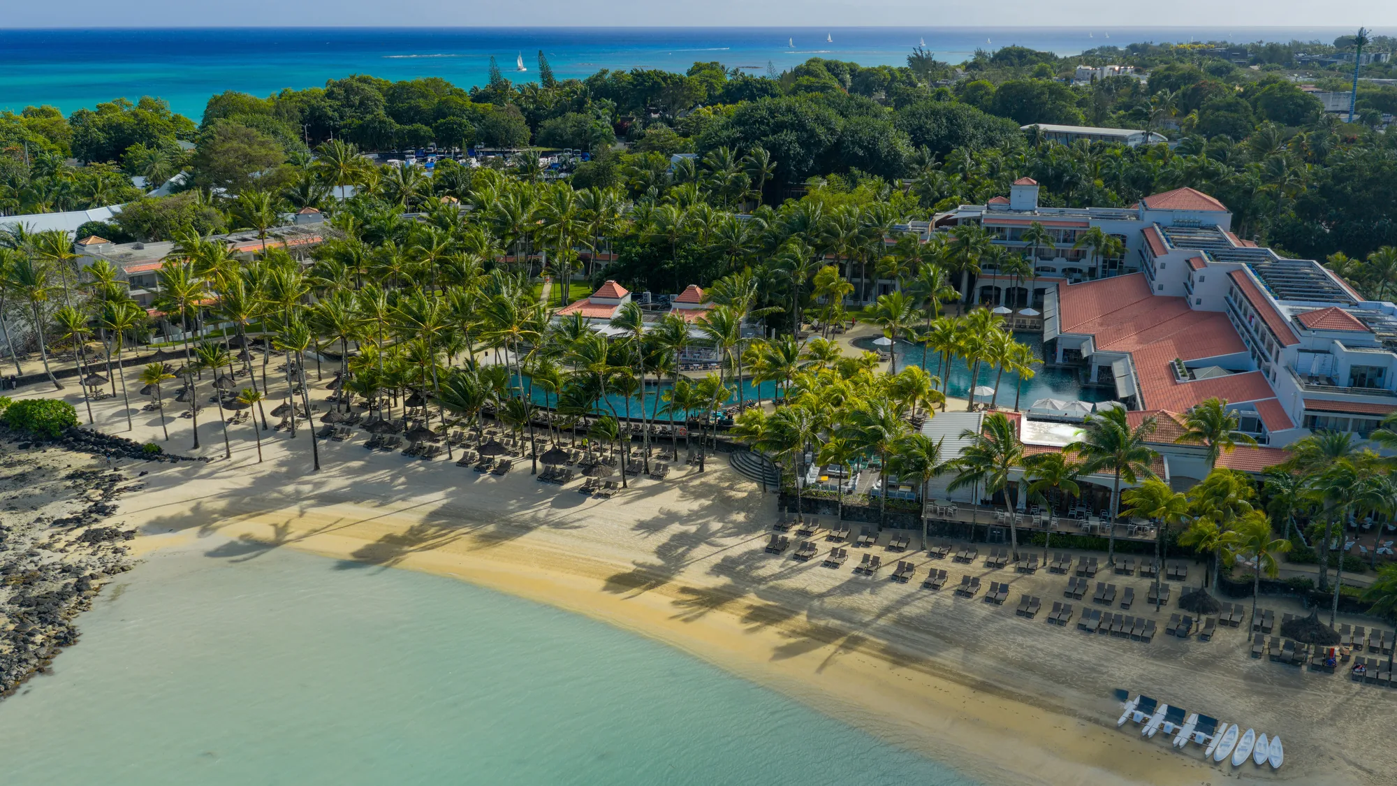 Aerial view of Mauricia Beachcomber, its red roofs and pools behind a curving beach on the bay
