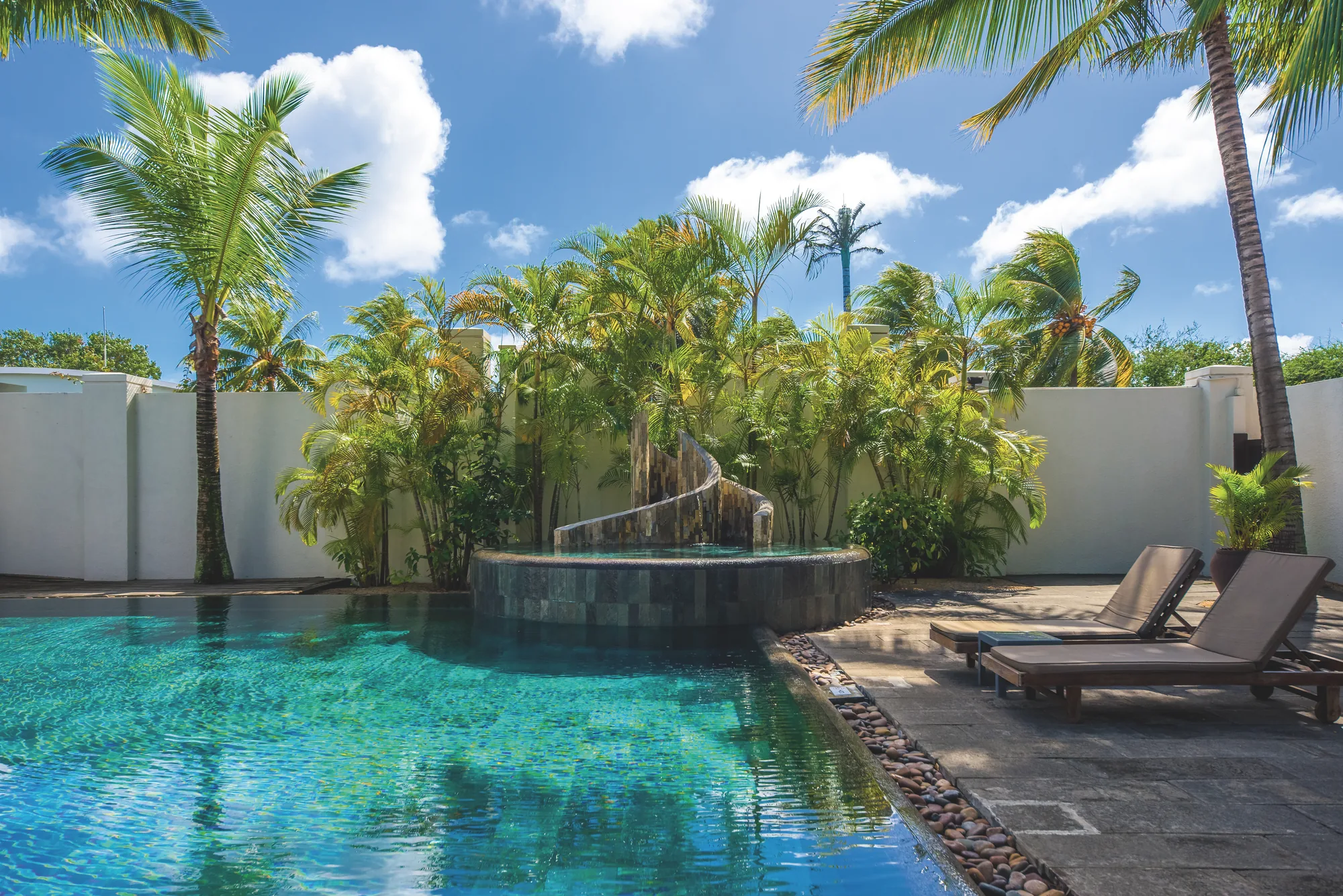 A pool with a rock waterfall and loungers, among palm trees