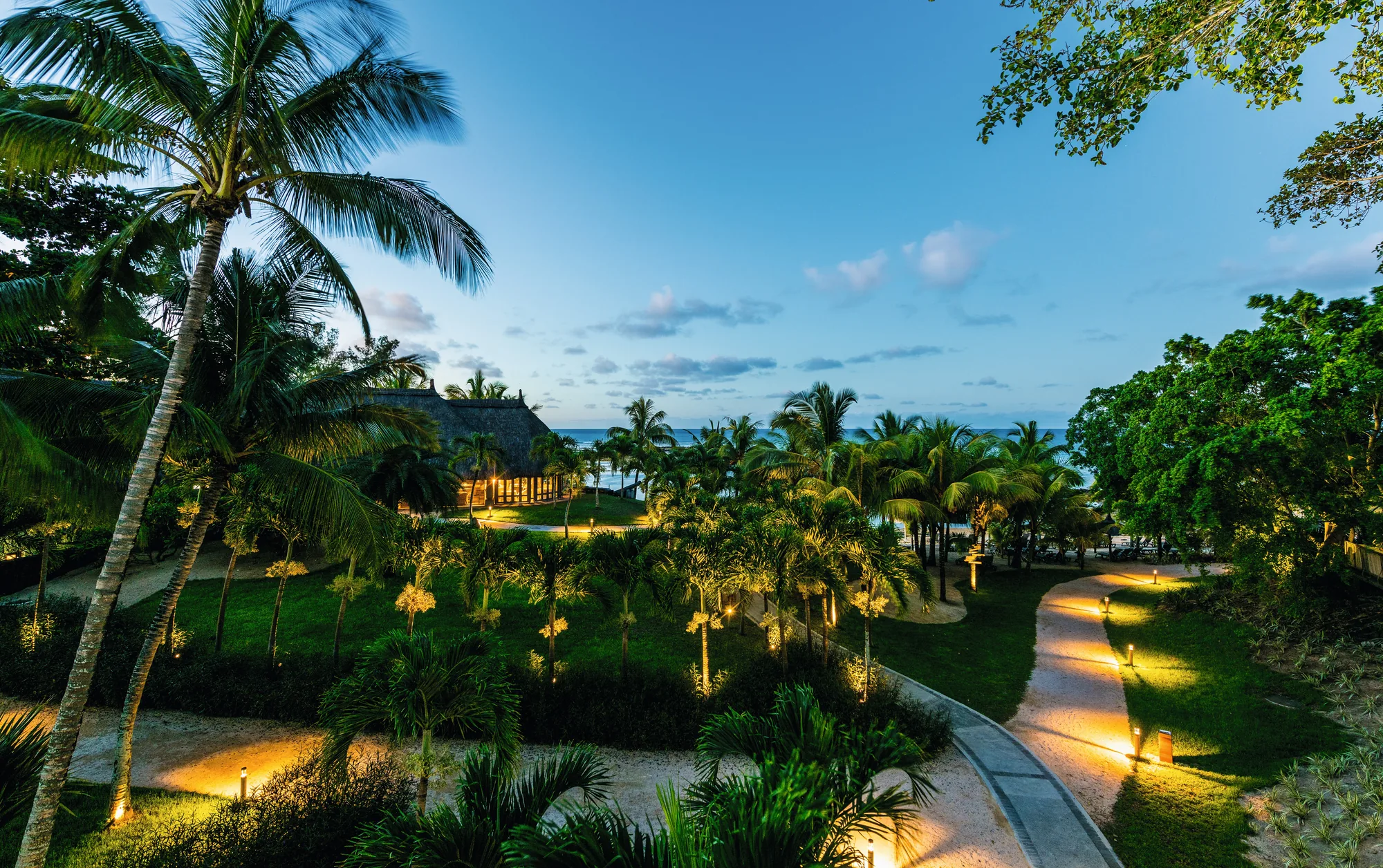 Garden paths lit at dusk among palm trees, with the sea beyond