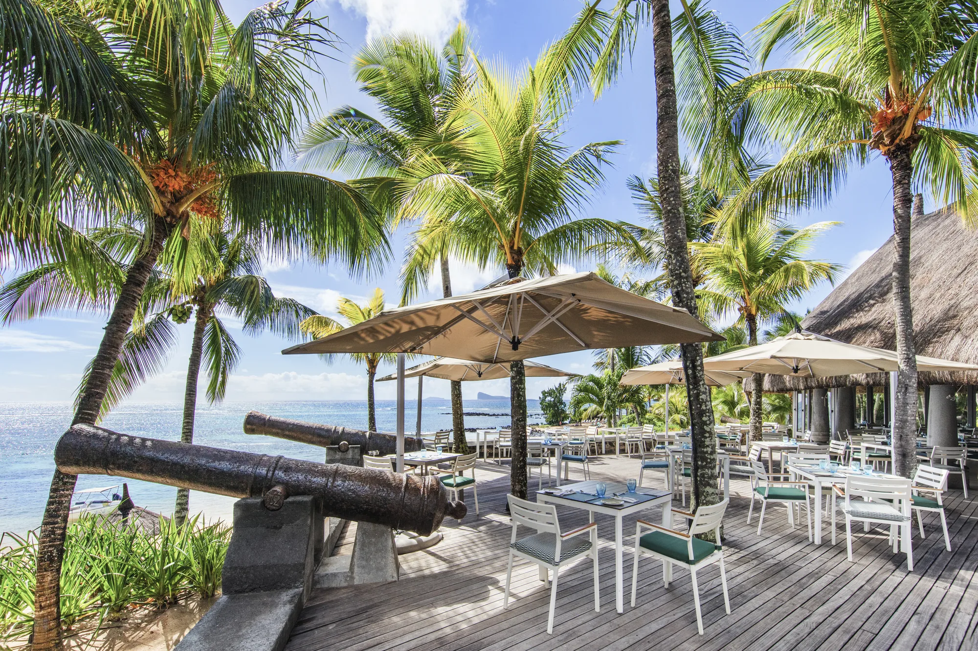 An old cannon beside a deck restaurant with parasols, overlooking the sea