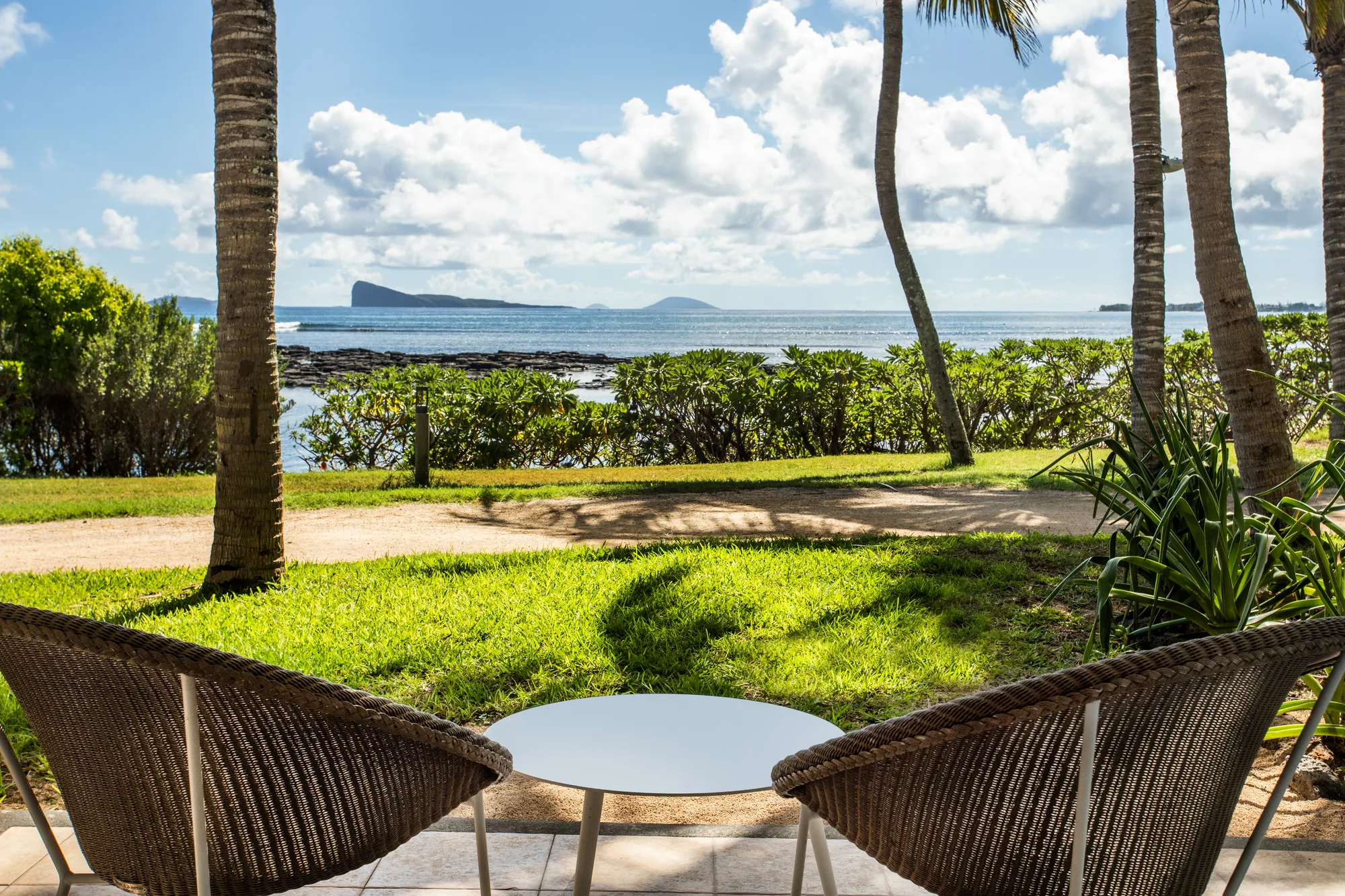 Two wicker chairs and a table on a lawn, looking out to the sea and an island offshore
