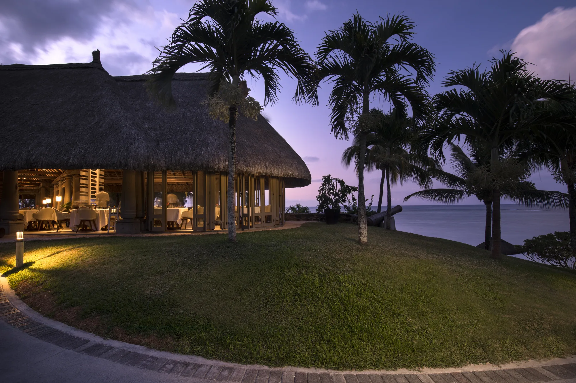 A thatched restaurant lit at dusk on a lawn beside the sea