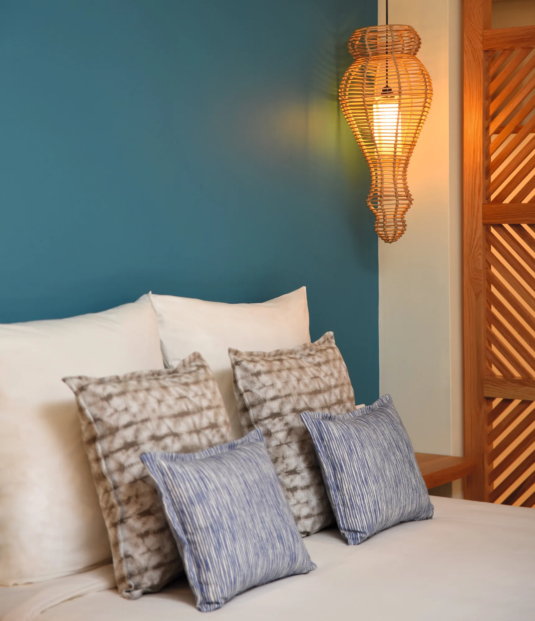 A bed with patterned cushions against a teal wall, lit by a woven lamp