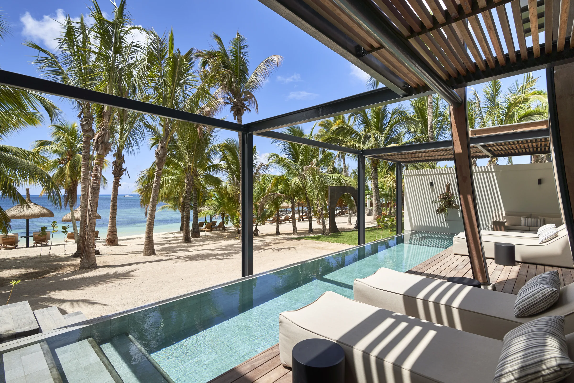 A pool terrace with daybeds under a slatted pergola, looking to the beach
