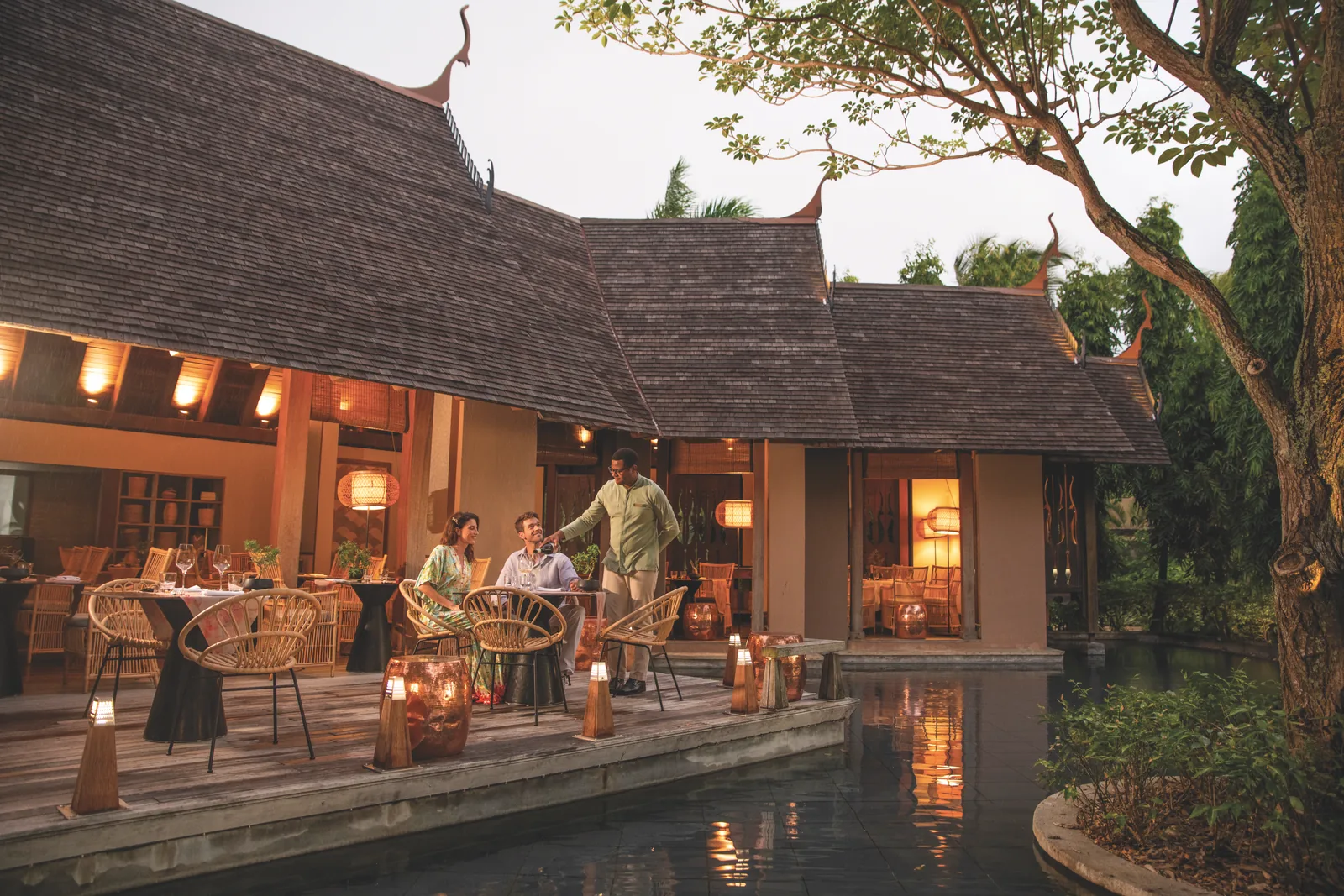Guests dining on a lit terrace beside a pond at dusk
