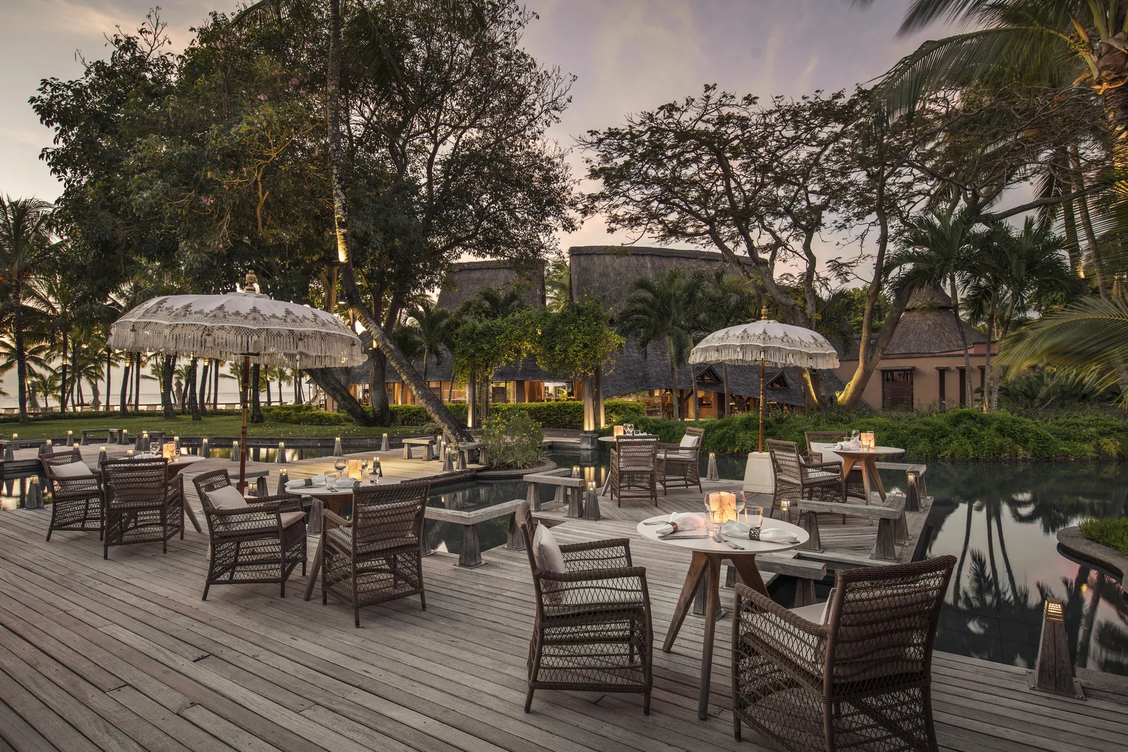 A deck restaurant with wicker chairs and parasols beside a pond at dusk