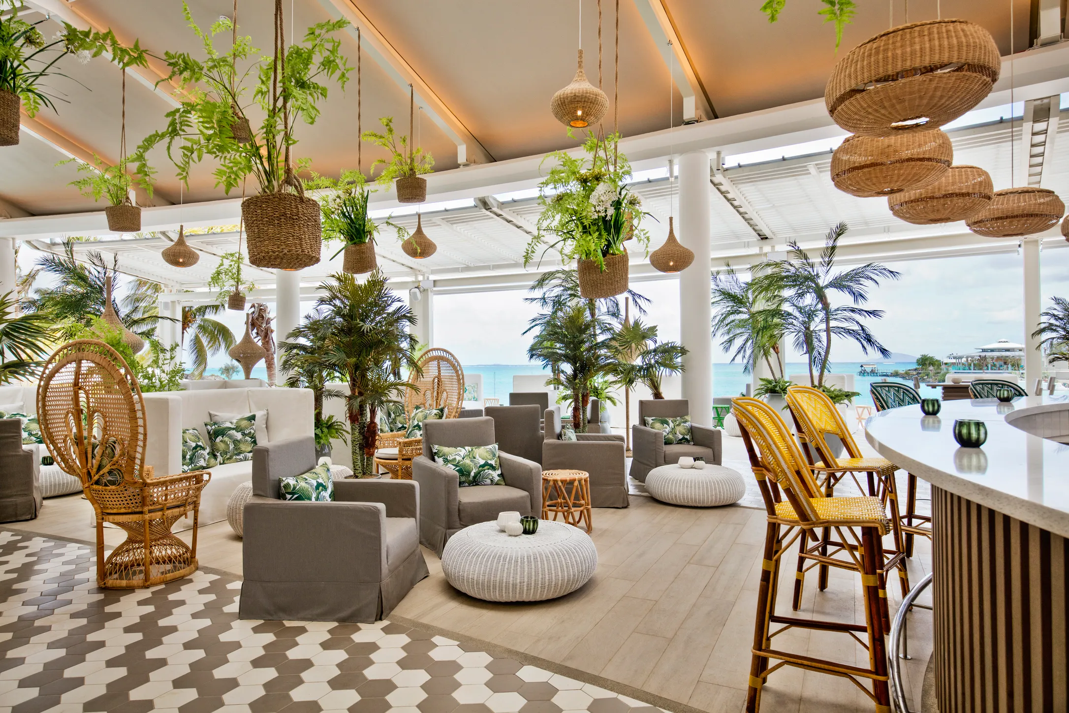 A lounge with hanging plants, rattan chairs and patterned floor tiles, open to the sea