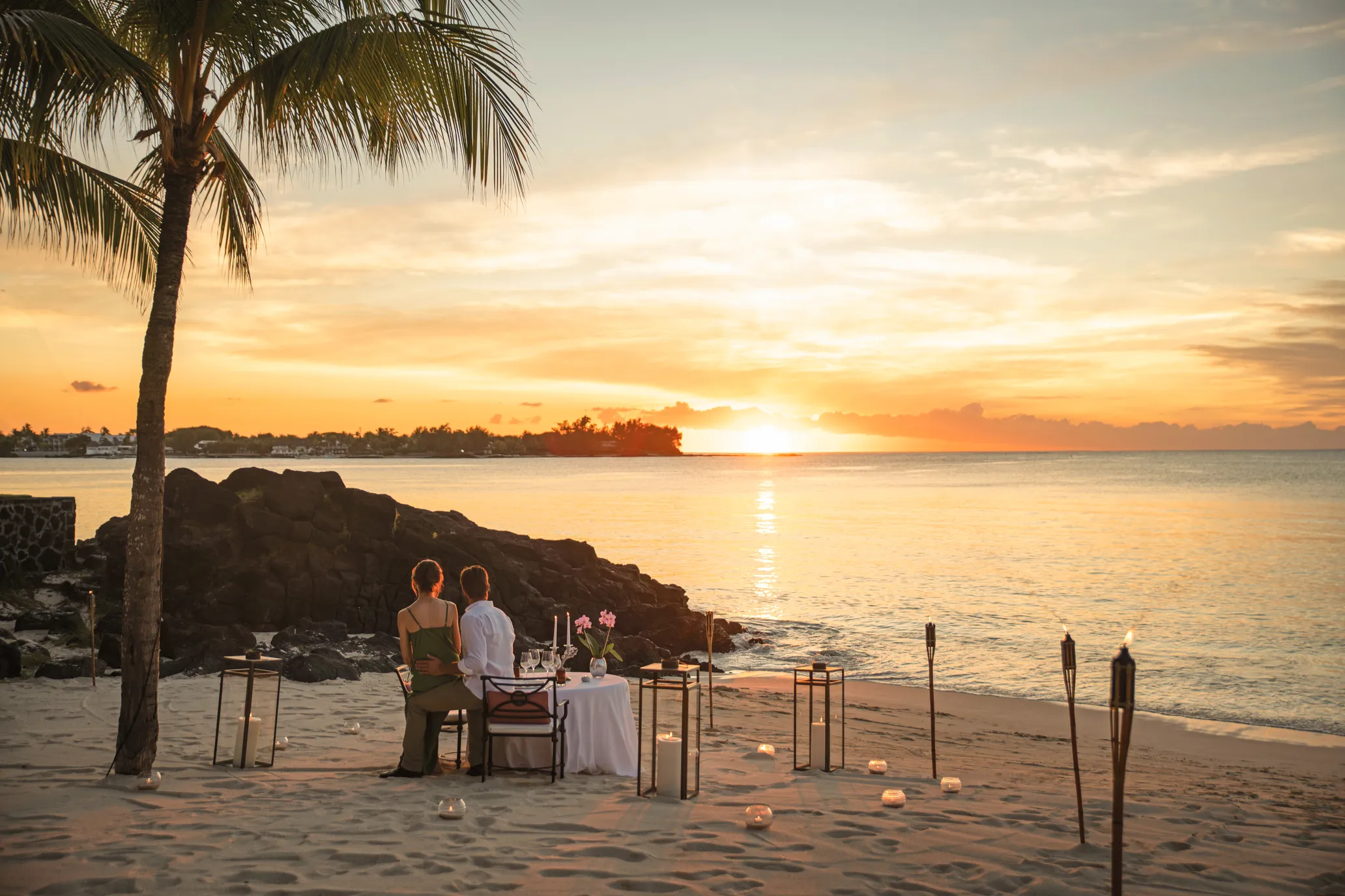 A couple dining on the beach at sunset, with lanterns set out on the sand