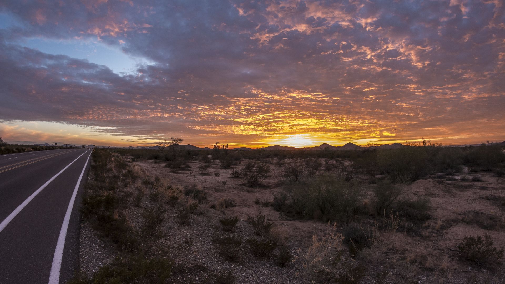 Two-lane road beside desert scrub at sunset, with orange clouds over low mountains on the horizon