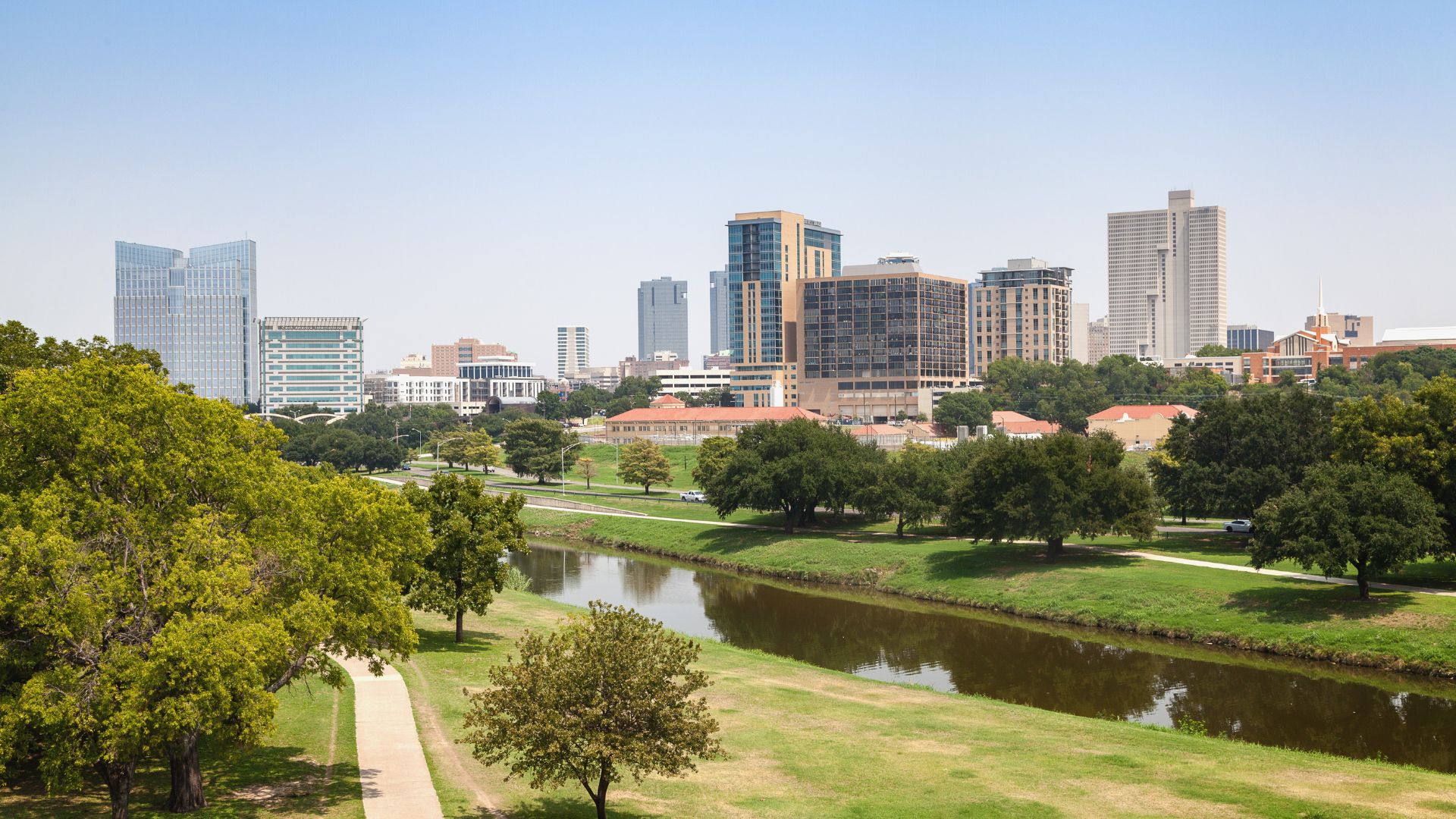 Downtown skyline rising beyond a tree-lined river and green parkland under a blue sky
