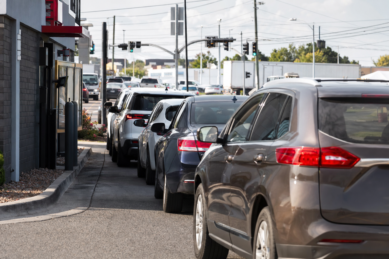 Busy fast food drive-thru lane with cars lined up, showing environmental challenges like road noise and acoustic interference, modern QSR restaurant exterior, multiple vehicles in queue, realistic daytime photography style, focus on the challenging acoustic environment with traffic and background activity, professional commercial photography