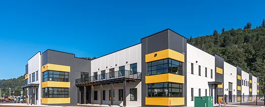 Modern industrial building with a white, gray, and yellow facade, featuring large windows and a surrounding forest backdrop.