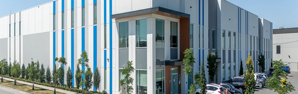 Large industrial office complex with a white and gray facade, accented by vertical blue stripes, landscaped with trees and shrubs along the sidewalk, and a row of parked cars in front.