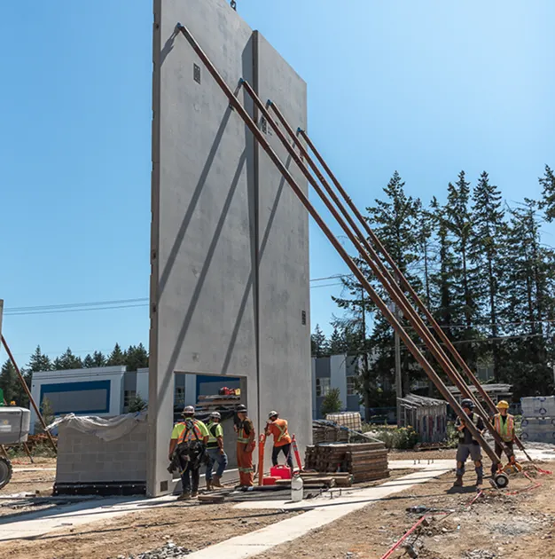 Construction workers securing a tall precast concrete wall panel with steel braces on a sunny day at a building site.