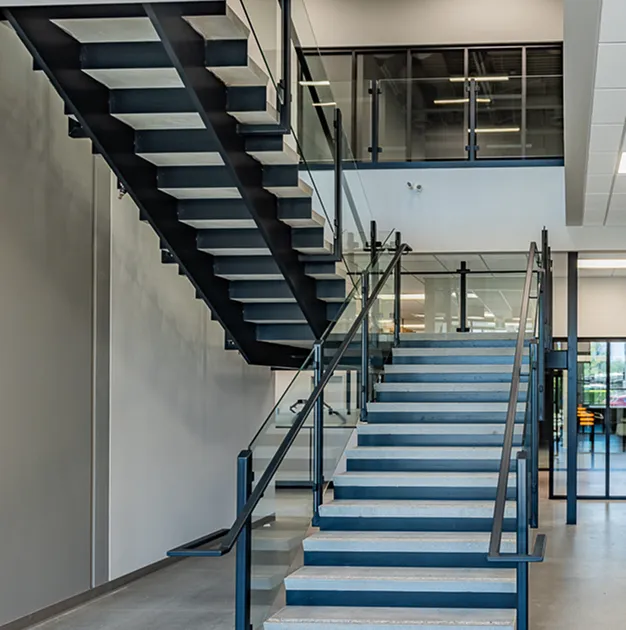 Interior view of a modern office building with a wide staircase, black metal framing, and glass railings leading to an upper level with enclosed offices.