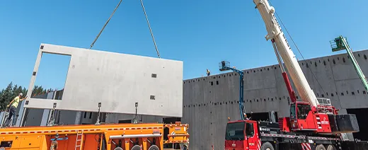 Construction site with a crane lifting a large precast concrete wall panel into place against a partially built structure under a clear blue sky.