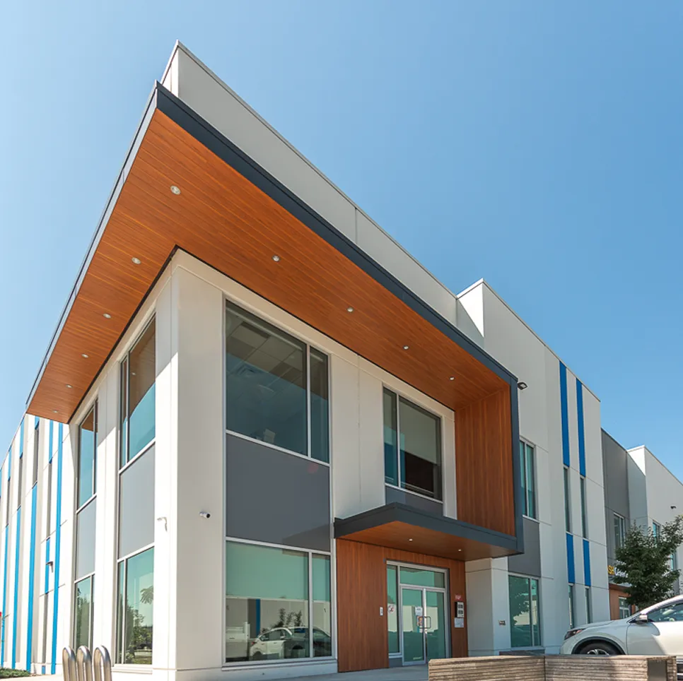 Contemporary two-story office building with large glass windows, gray and white exterior panels, and warm wood accents under the overhang, set against a clear blue sky.