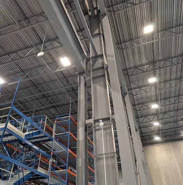 Interior of an industrial facility showing tall steel structural supports, overhead lighting, and multi-level blue and orange storage racks.