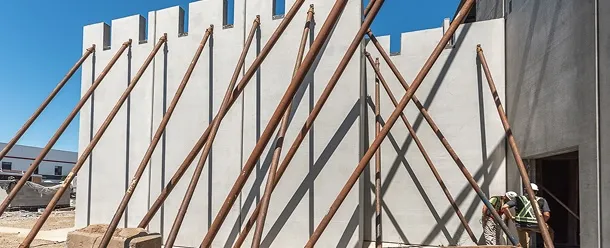 Construction site with tall precast concrete walls supported by multiple diagonal steel braces, with workers standing near the structure.