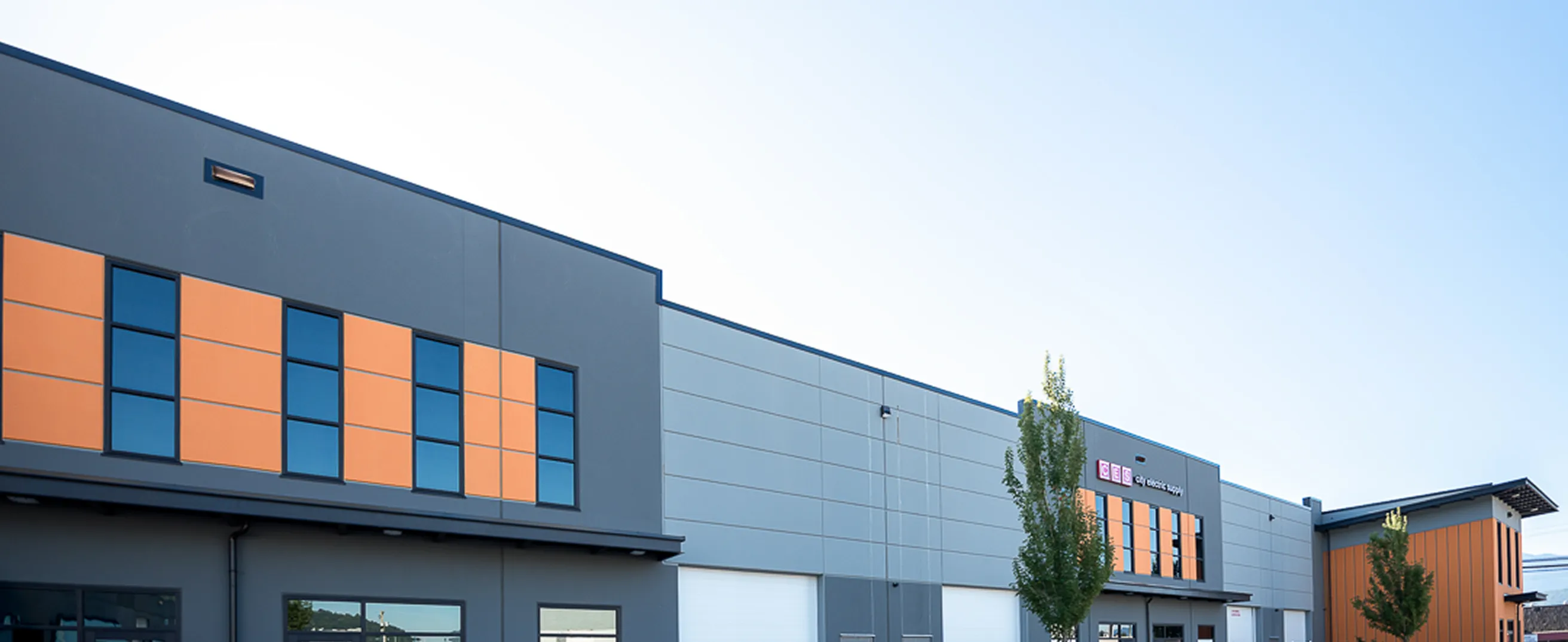 Industrial building with a gray facade, orange accent panels, large windows, and multiple loading bay doors under a clear sky.