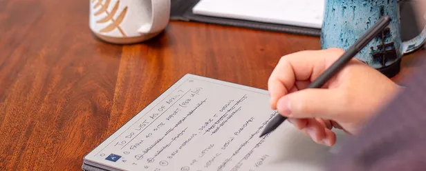 Close-up of a person writing a to-do list on a tablet with a stylus, next to a blue ceramic mug on a wooden table.