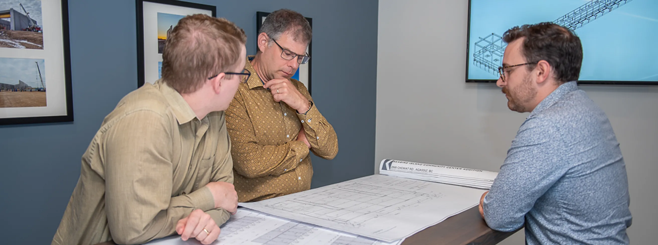 Three men in an office meeting, discussing architectural blueprints on a table, with framed construction photos and a digital design display in the background.