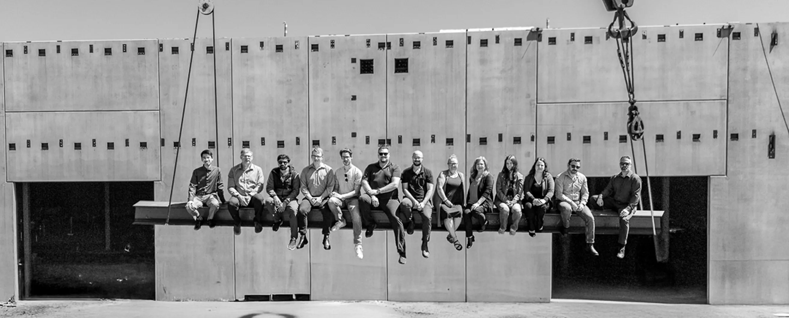 Black-and-white photo of a group of people sitting on a suspended steel beam in front of a large concrete wall at a construction site.