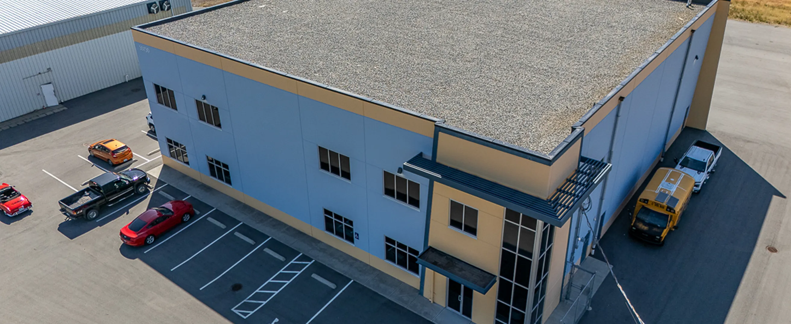 Aerial view of a two-story industrial building with a blue and beige facade, surrounded by parked cars, including a yellow school bus and red vehicles.