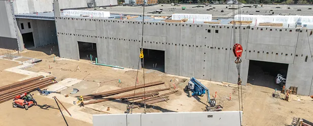 Construction site with a large partially built concrete structure, open bays, cranes, and scattered steel beams under a clear sky.