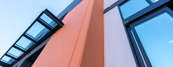Construction site with a large crane lifting a precast concrete wall panel into position, supported by an orange flatbed trailer under a clear blue sky.