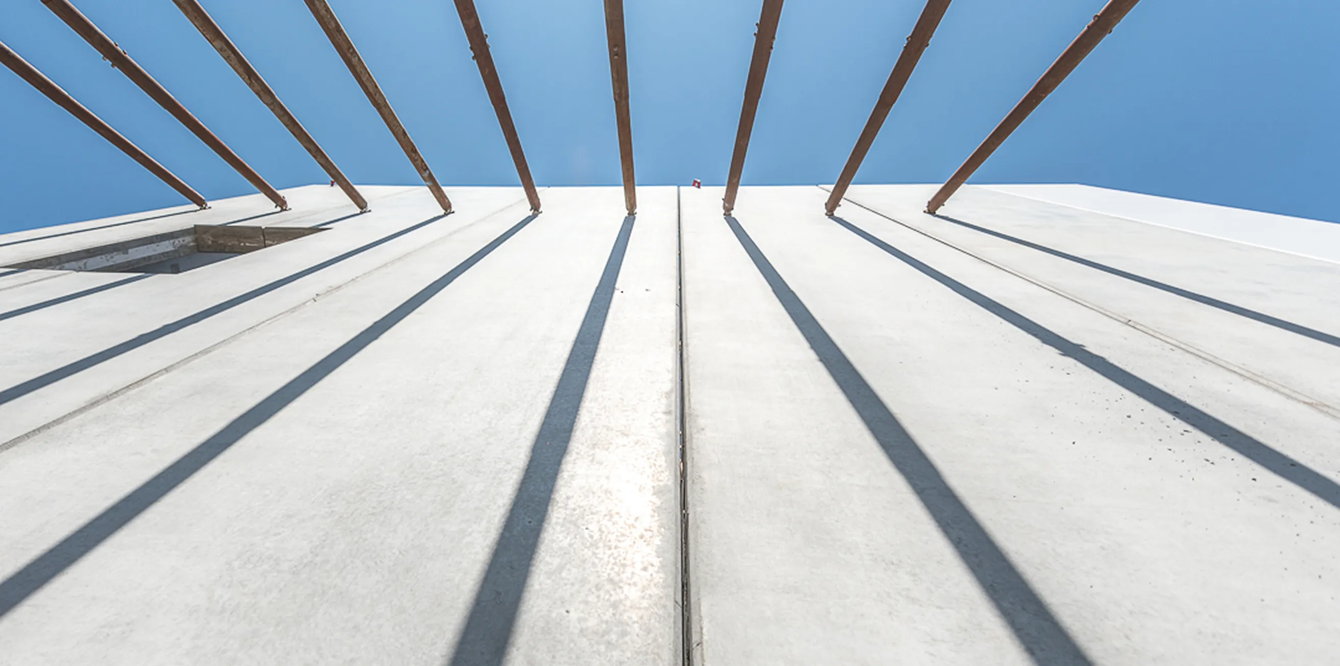 Upward view of a precast concrete wall with diagonal steel braces casting long shadows against a bright blue sky.