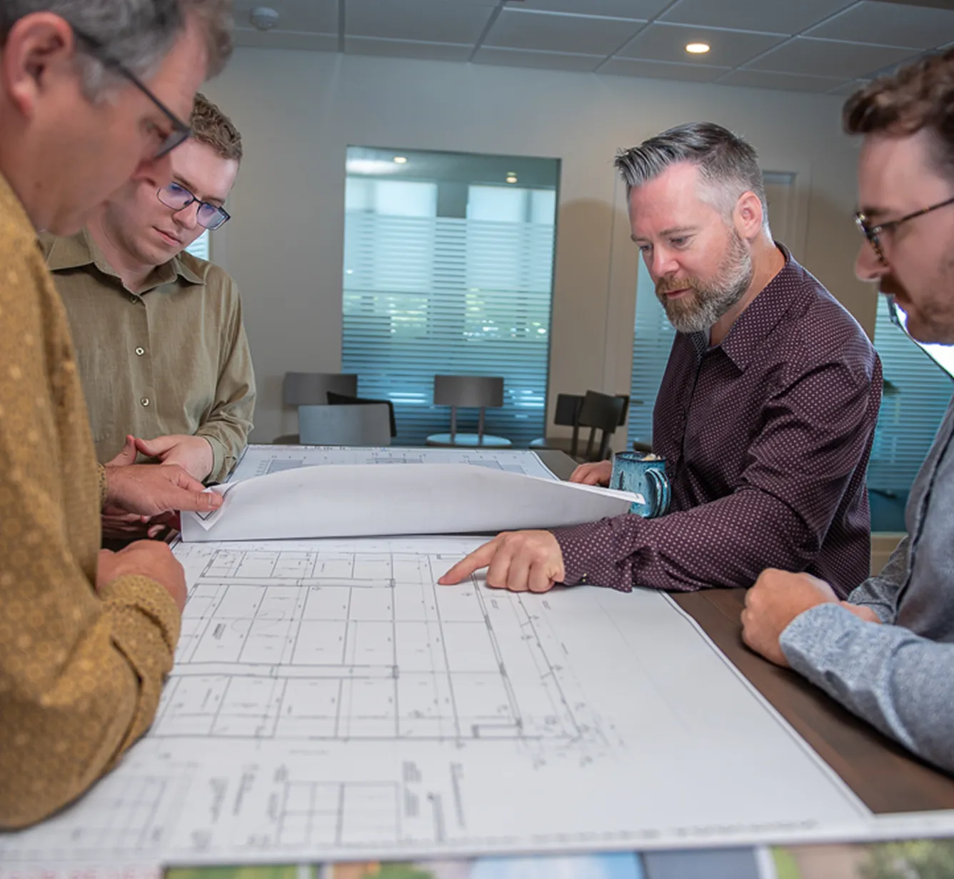 Group of four engineers reviewing architectural blueprints at a table, with one pointing at specific details on the plans.