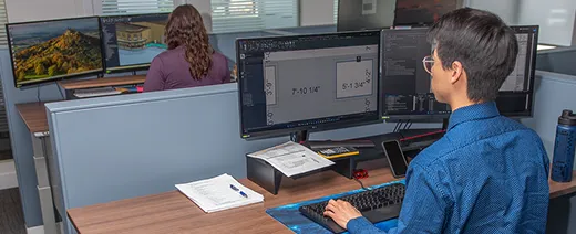 Office scene with two employees working at dual-monitor setups, one focused on technical drawings displayed on the screen.