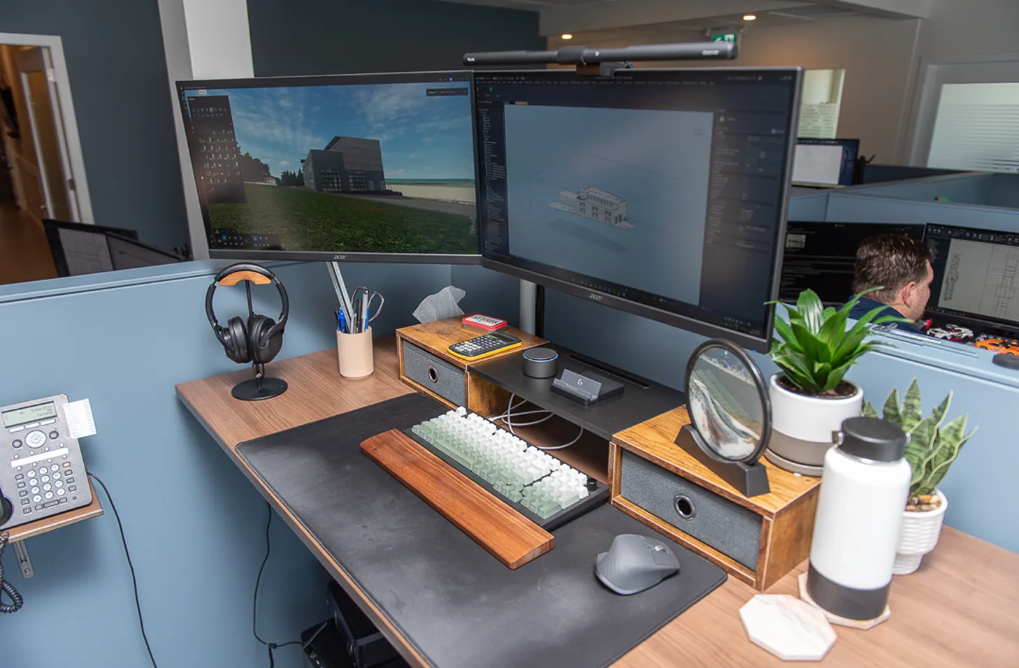 Organized office workstation with dual monitors displaying architectural design software, a wooden keyboard tray, plants, and personal desk accessories.