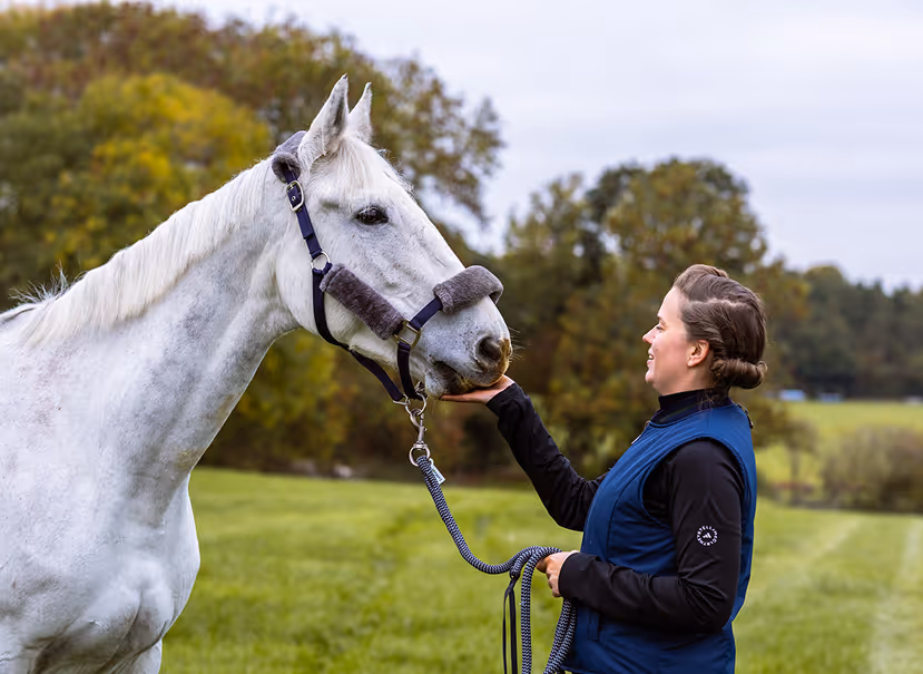 Woman smiling and gently feeding a white horse in a green outdoor field with trees in the background.