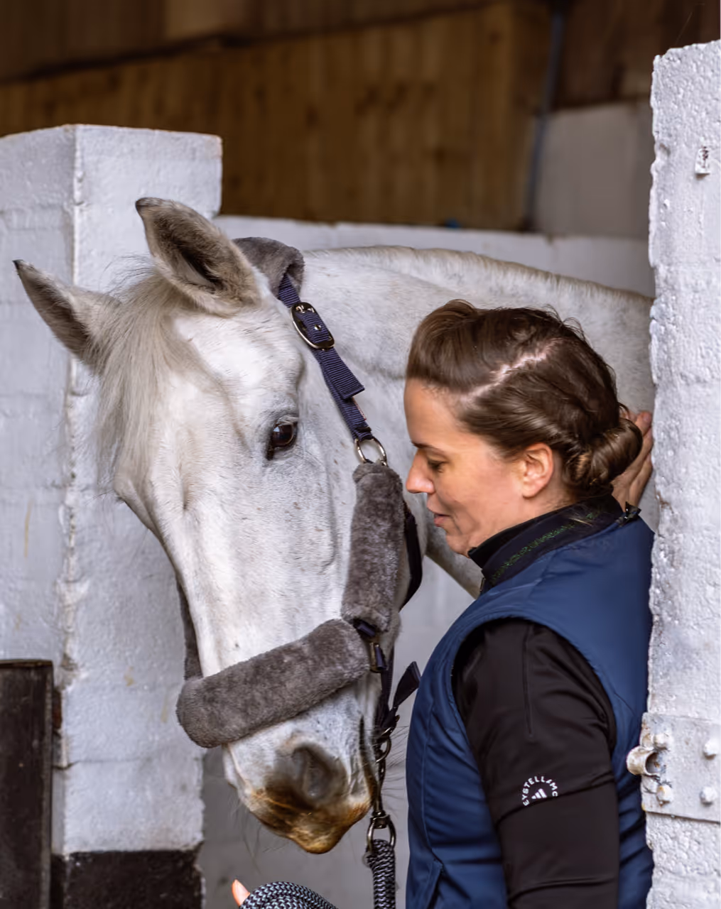 Woman standing close and gently touching the head of a white horse inside a stable.