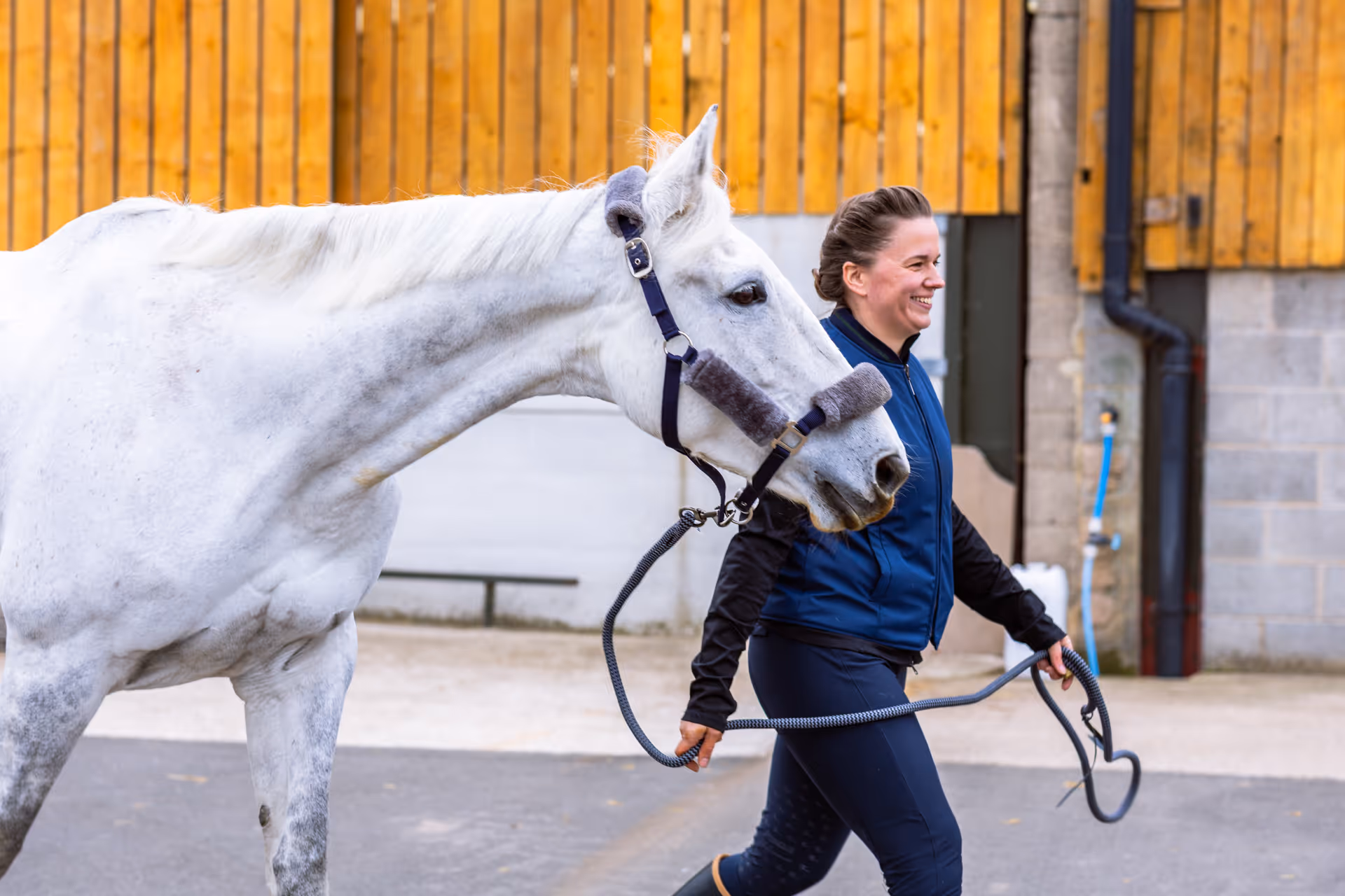 Smiling woman in a vest jacket leading a white horse with a halter outside near wooden and concrete buildings.
