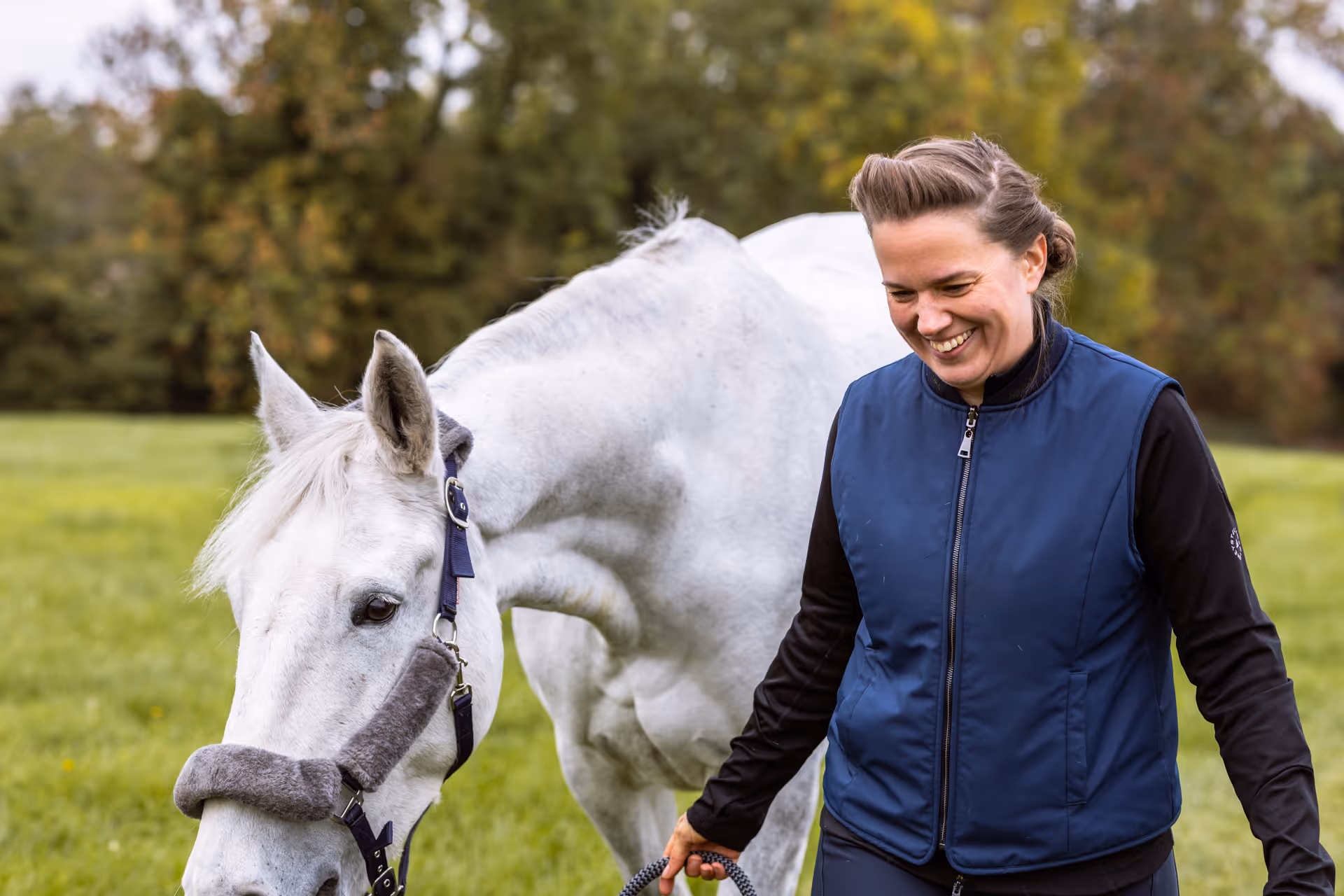 Smiling woman in a navy vest walking a white horse on a grassy field with trees in the background.