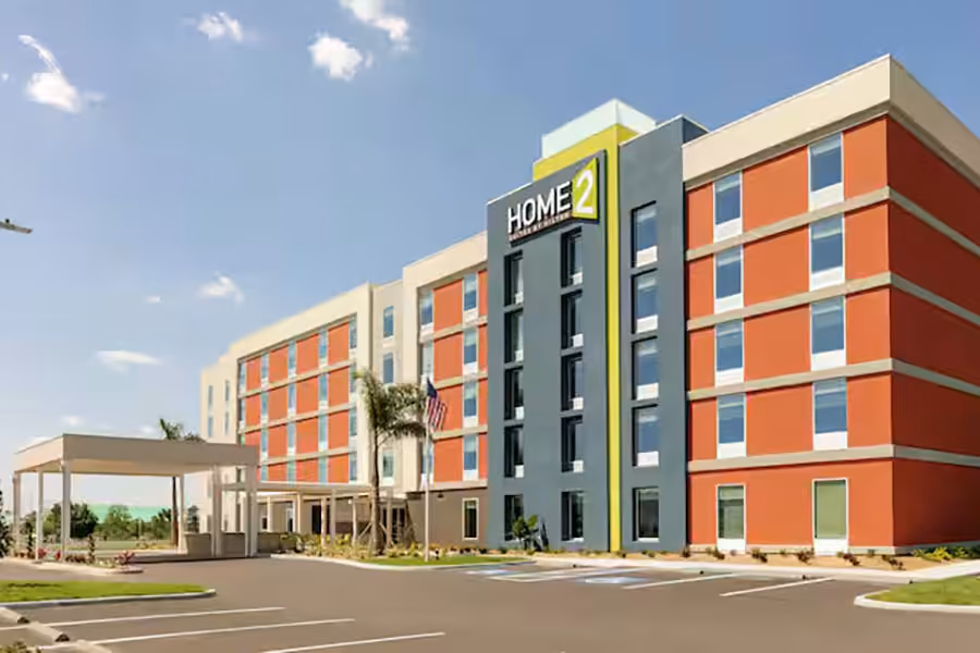 Four-story Home2 Suites hotel with red and beige exterior, front entrance canopy, and empty parking lot.