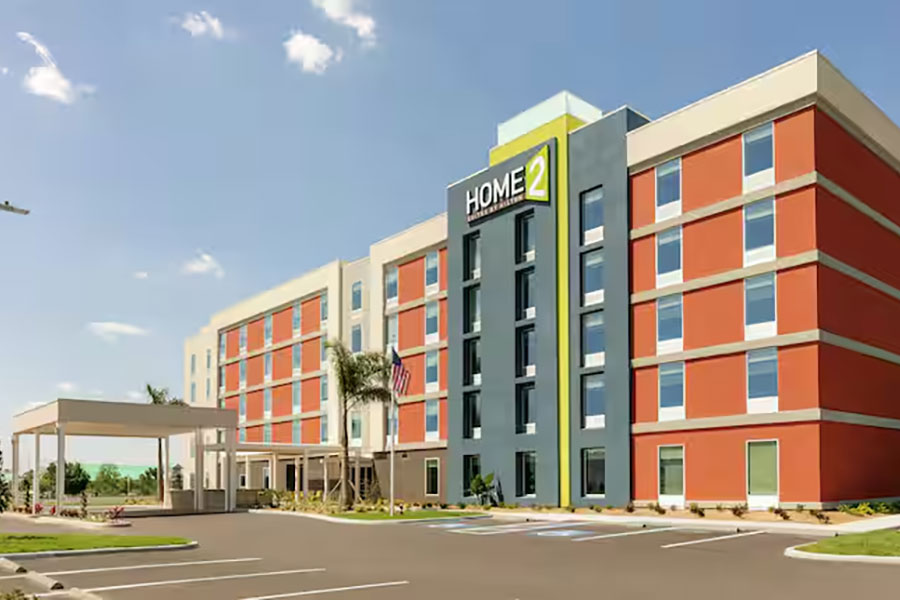 Four-story Home2 Suites hotel with red and beige exterior, front entrance canopy, and empty parking lot.