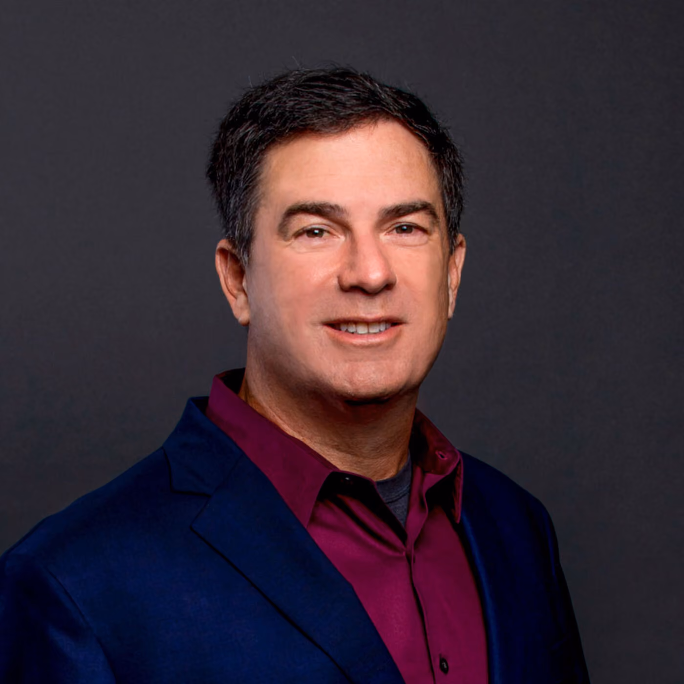 Man in a dark blue suit and maroon shirt poses against a plain dark background, facing the camera and smiling.