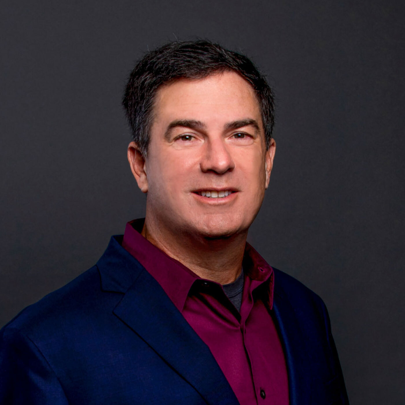 Man in a dark blue suit and maroon shirt poses against a plain dark background, facing the camera and smiling.