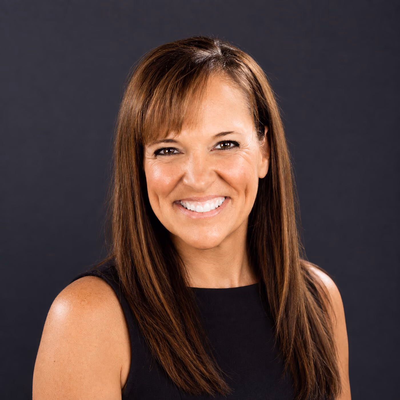 Woman with straight brown hair wearing a sleeveless black top, smiling in front of a plain dark background.