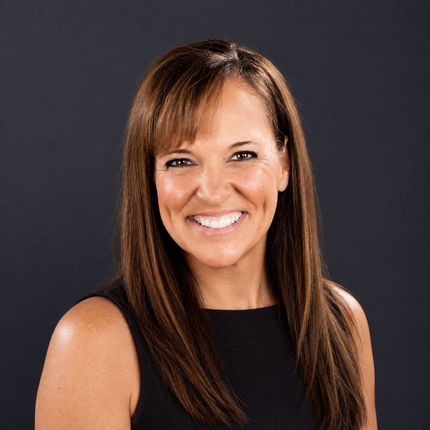 Woman with straight brown hair wearing a sleeveless black top, smiling in front of a plain dark background.