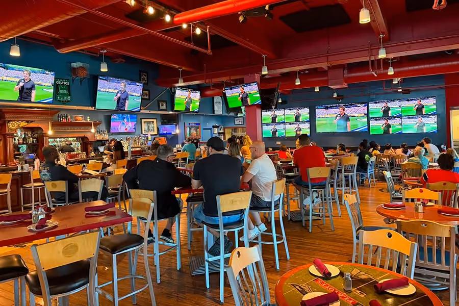 People sit at tables in a sports bar watching multiple TVs showing a soccer game, with drinks on the tables.