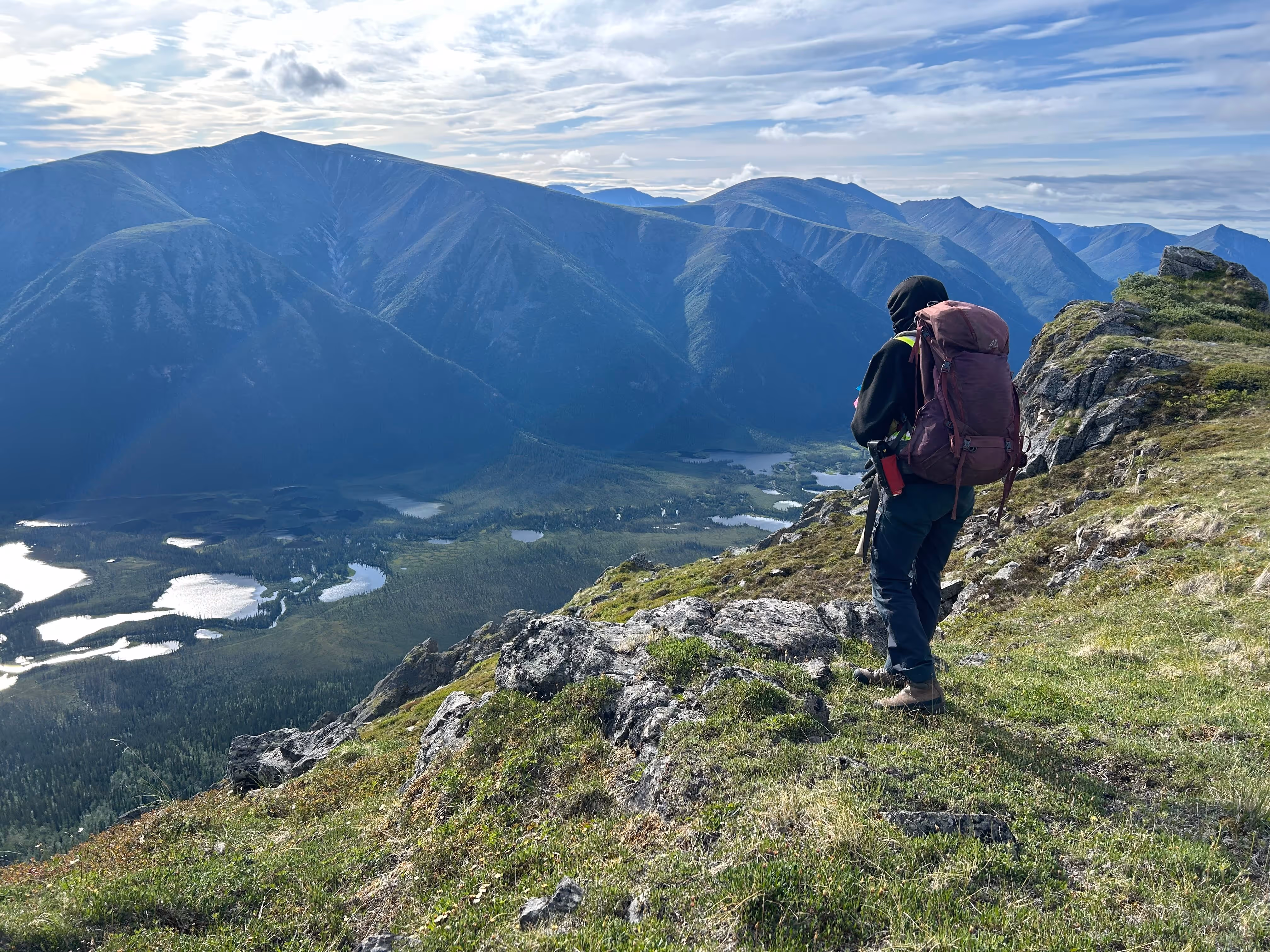 Man with Backpack on hill looking down below and mountains on background