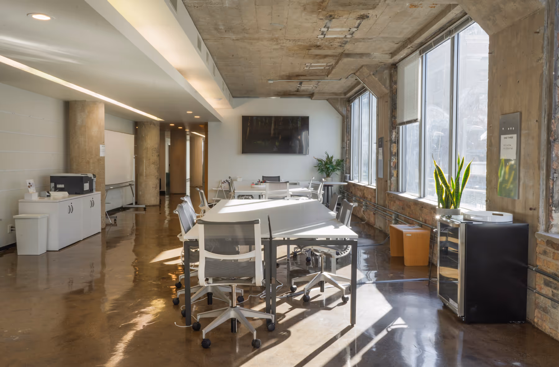 Bright modern conference room with white tables, rolling mesh chairs, large windows, and concrete ceiling and walls.