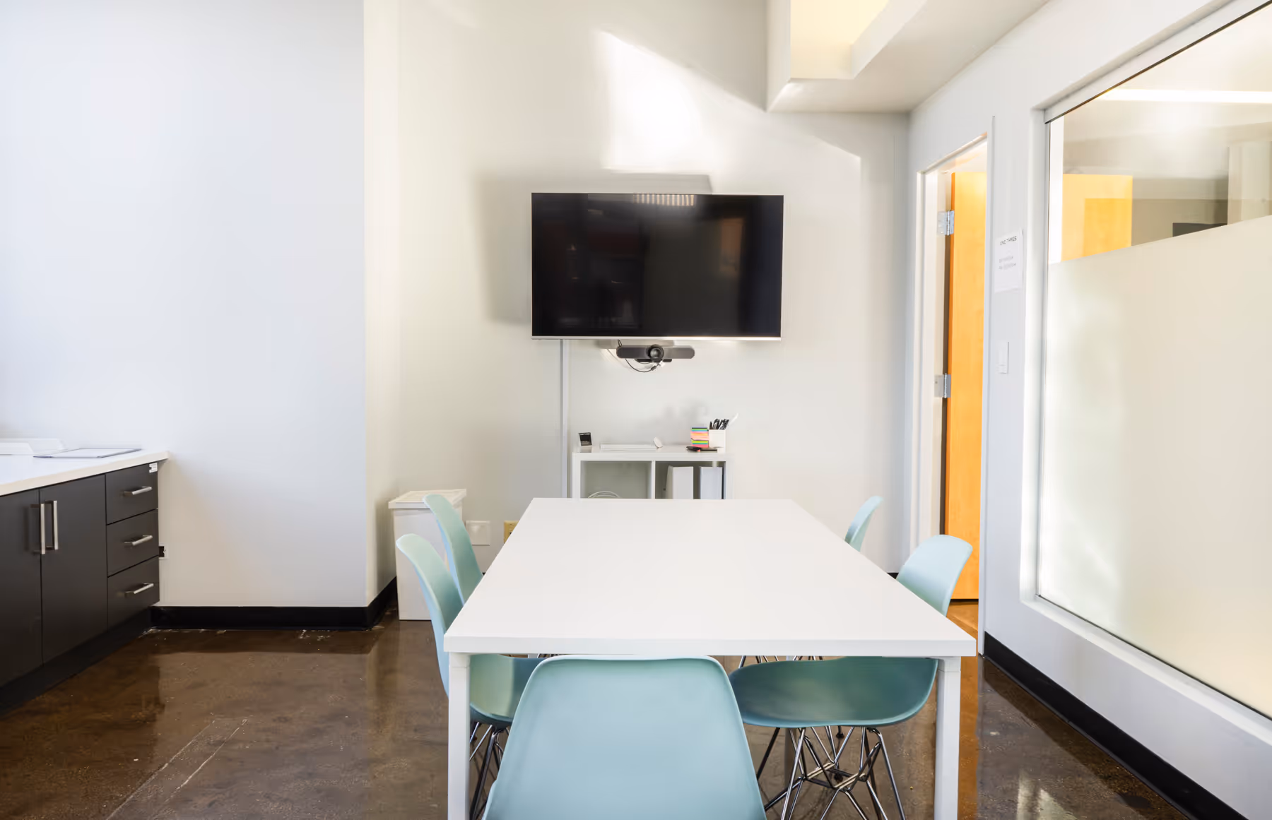 Modern conference room with a white rectangular table surrounded by six light blue chairs, a wall-mounted TV, and a small white shelf with office supplies.