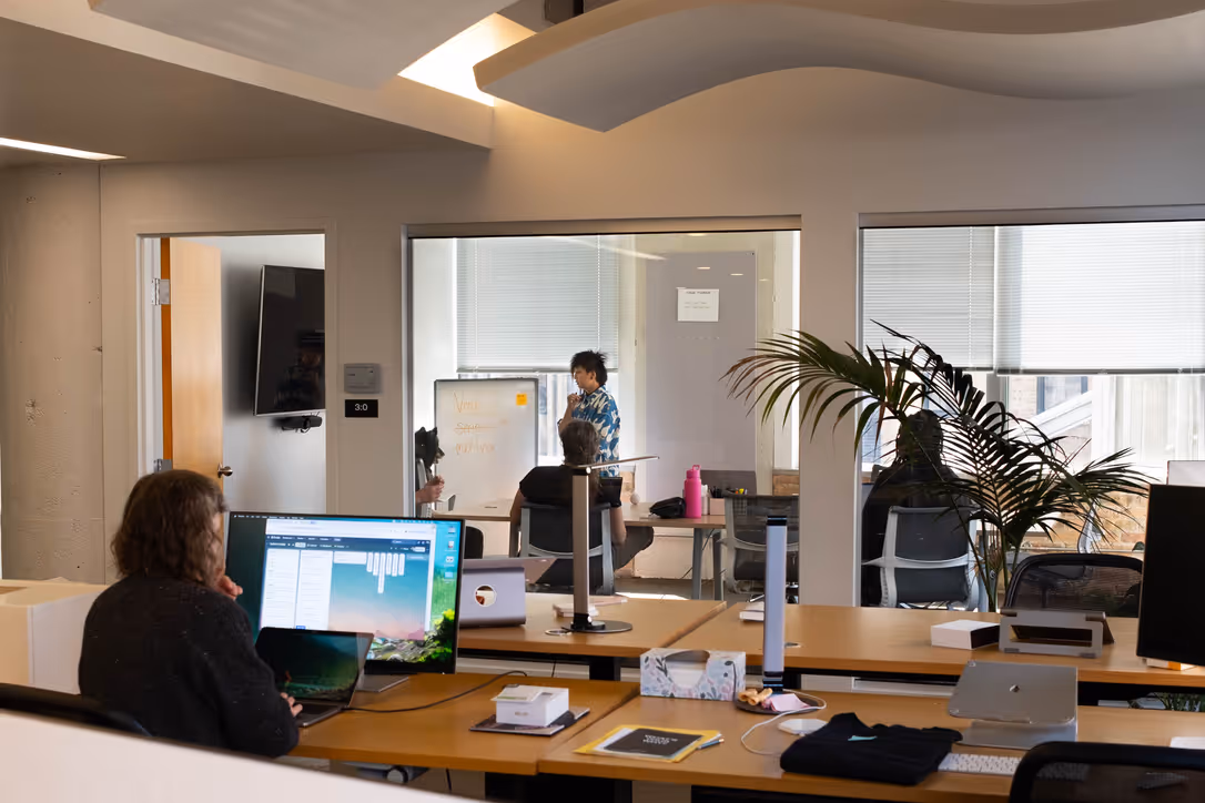 Office workspace with a person working at a desk and a meeting with three people discussing at a whiteboard in a glass-walled conference room.