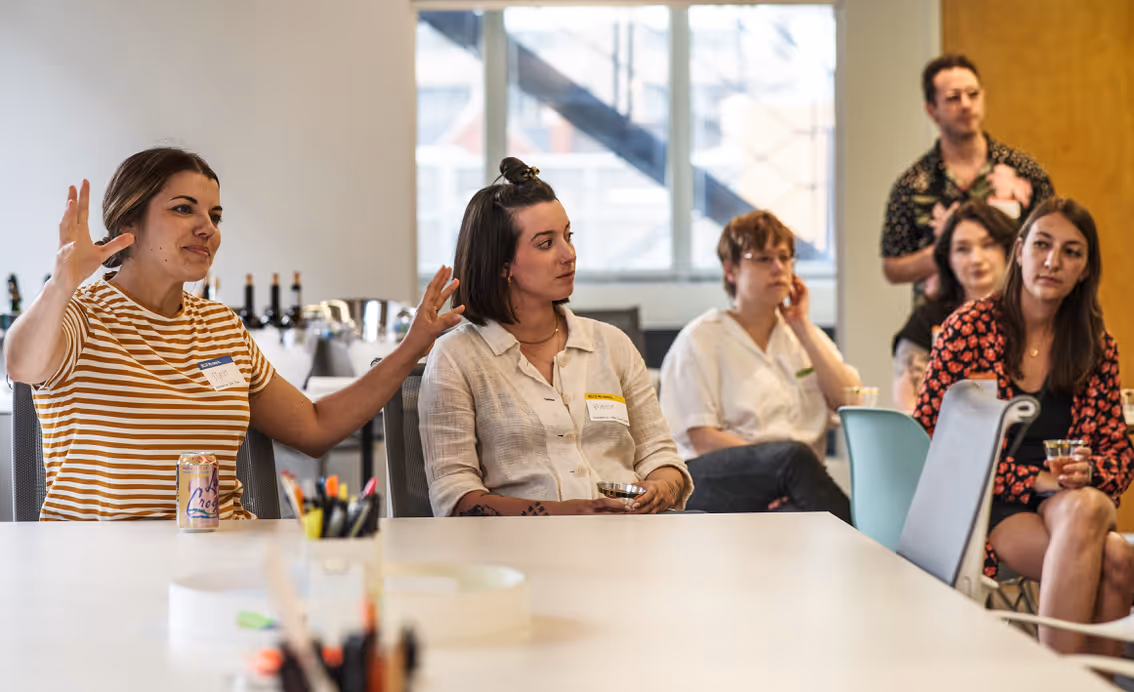 Group of six people seated around a conference table, one woman in striped shirt speaking with hands raised.